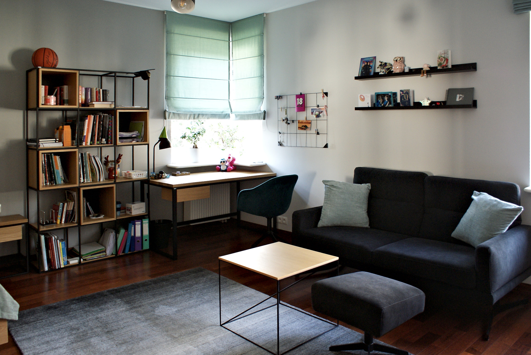 Pokój dzienny z drewnianą podłogą, szarą sofą, biurkiem przy oknie z roletą rzymską, regałem z książkami i stolikiem kawowym na szarym dywanie.