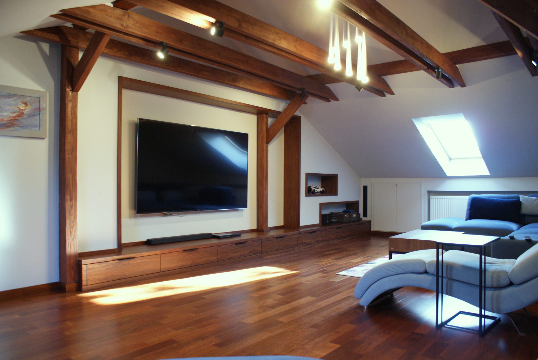 Salon na poddaszu z drewnianą konstrukcją stropu, zabudową TV z szafkami, szarą sofą i oknem dachowym.