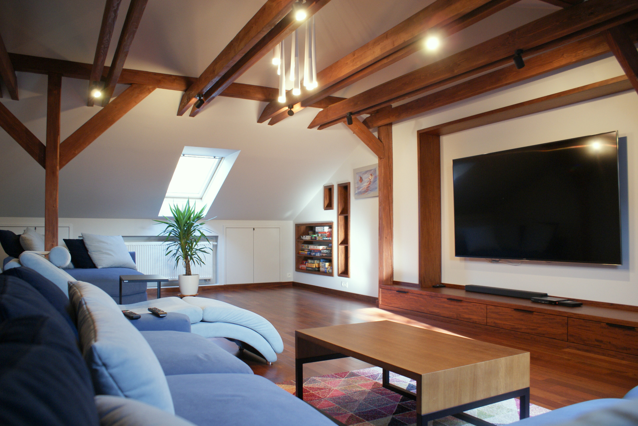 Jasny salon na poddaszu z drewnianymi belkami, niebieską sofą modułową, telewizorem w drewnianej obudowie i oknem dachowym z rośliną doniczkową.