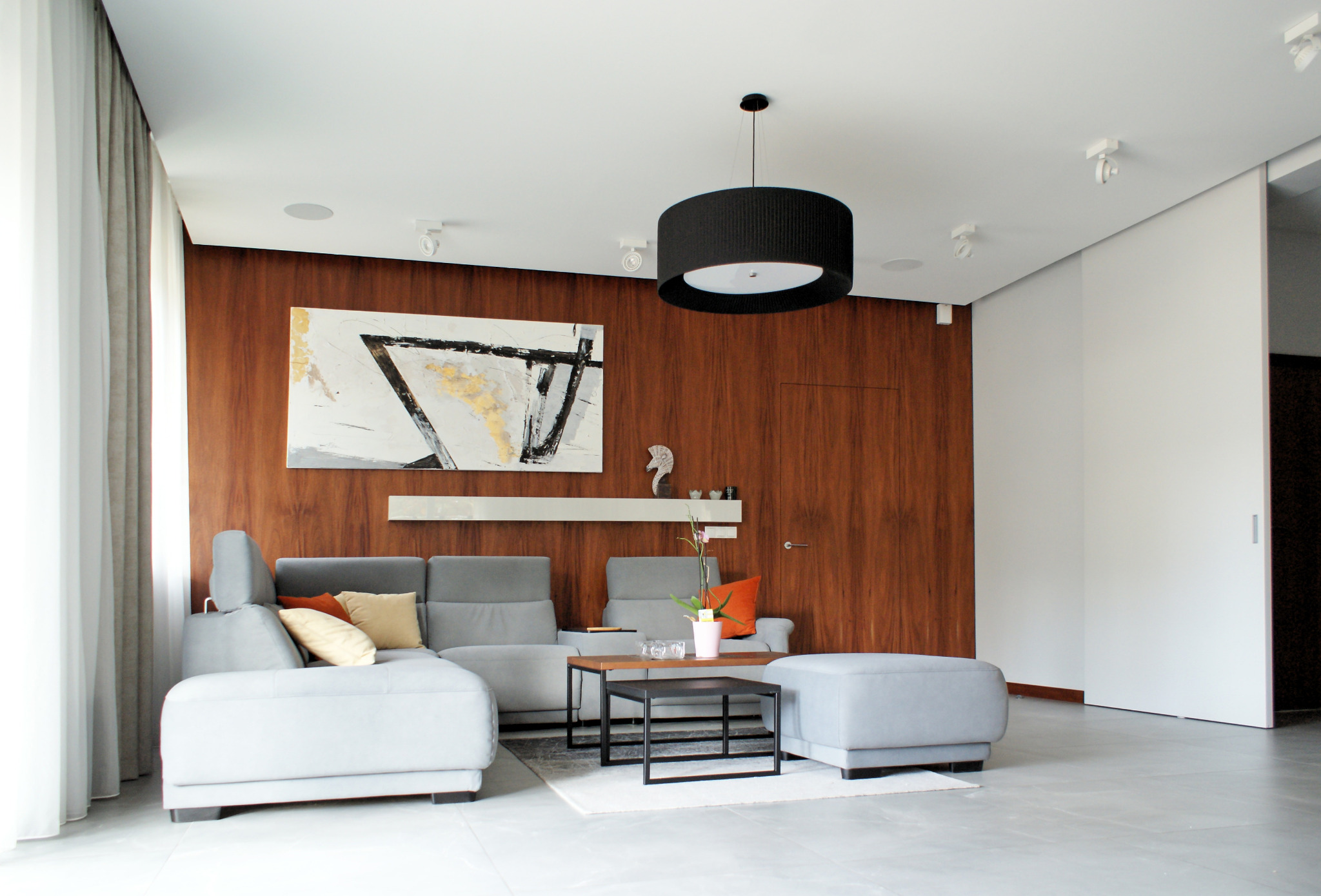 Nowoczesny salon z szarą sofą narożną, drewnianą ścianą z abstrakcyjnym obrazem, czarnym okrągłym żyrandolem i betonową podłogą.