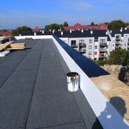 Nowy, płaski dach pokryty papą termozgrzewalną z widocznym wiadrem po smole i przygotowanymi deskami na tle budynków mieszkalnych.