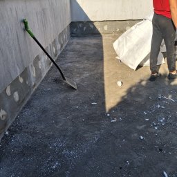 Flat Roof Pawel Canert - Przygotowanie do izolacji fundamentów: oczyszczona powierzchnia, łopata oparta o ścianę, pracownik z workiem materiału izolacyjnego w tle.