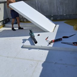 Flat Roof Pawel Canert - Montaż termoizolacji dachu płaskiego: białe płyty styropianowe układane na żółtej folii, widoczne narzędzia i fragment sylwetki pracownika. Jasne, słoneczne oświetlenie.