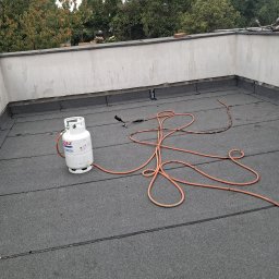 Flat Roof Pawel Canert - Prace dekarskie na płaskim dachu: biała butla gazowa, palnik dekarski z pomarańczowym wężem. W tle fragment budynku i zieleń drzew. Widoczny dekarz przy pracy.