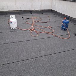 Flat Roof Pawel Canert - Praca na dachu płaskim: dekarz z palnikiem i butlami gazowymi na tle zieleni. Widoczna papa dachowa i narzędzia. Ujęcie z góry, podkreślające detale wykonania.