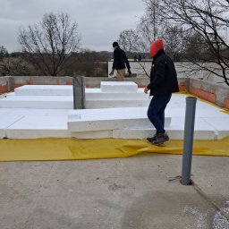 Flat Roof Pawel Canert - Prace izolacyjne na dachu: białe płyty styropianowe ułożone na żółtej folii, dwóch pracowników w tle. Widoczne elementy konstrukcyjne dachu i otoczenie.