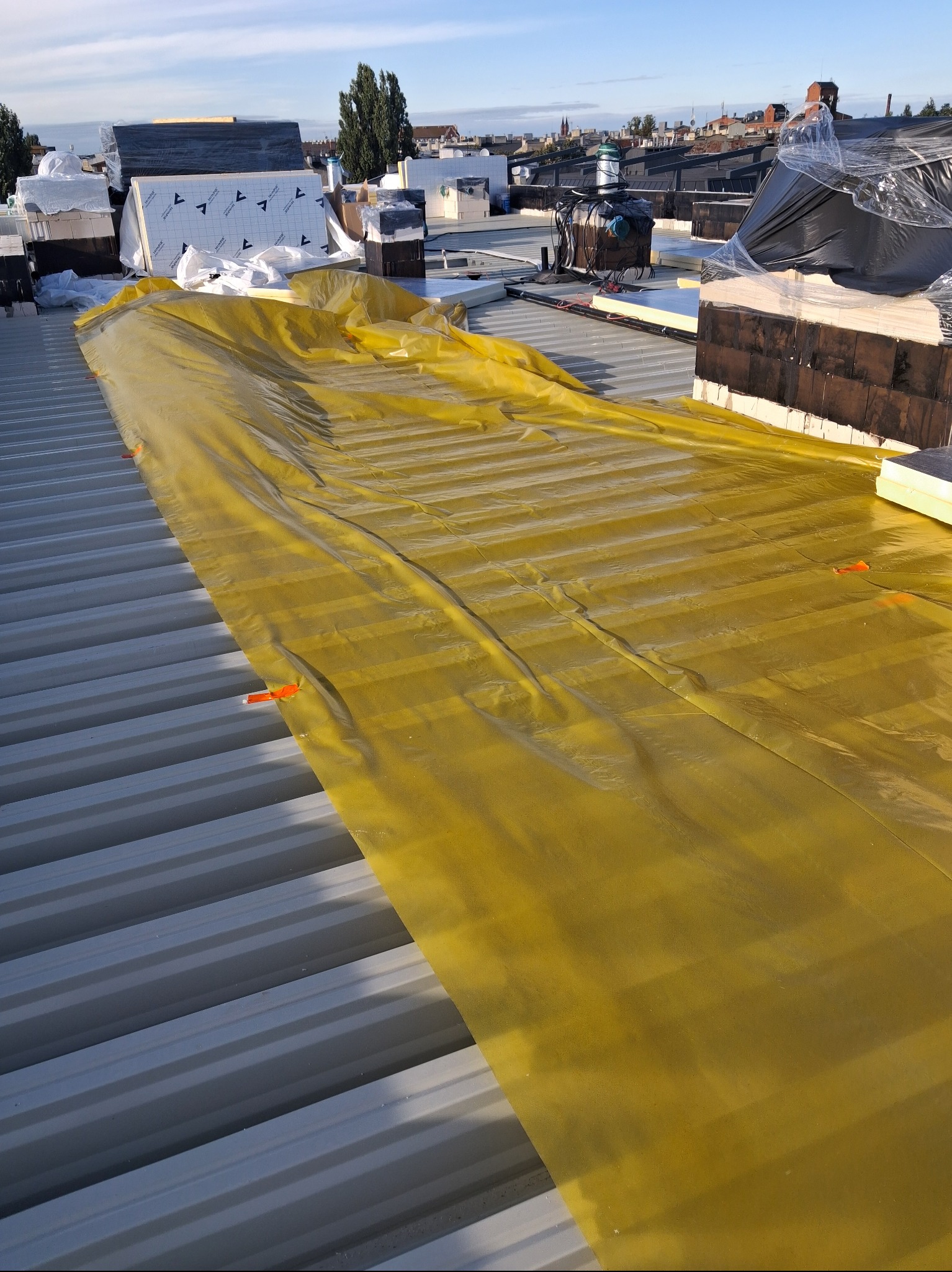 Żółta membrana dachowa rozłożona na metalowym dachu z widocznymi elementami izolacji i panoramą miasta w tle, w słoneczny dzień.
