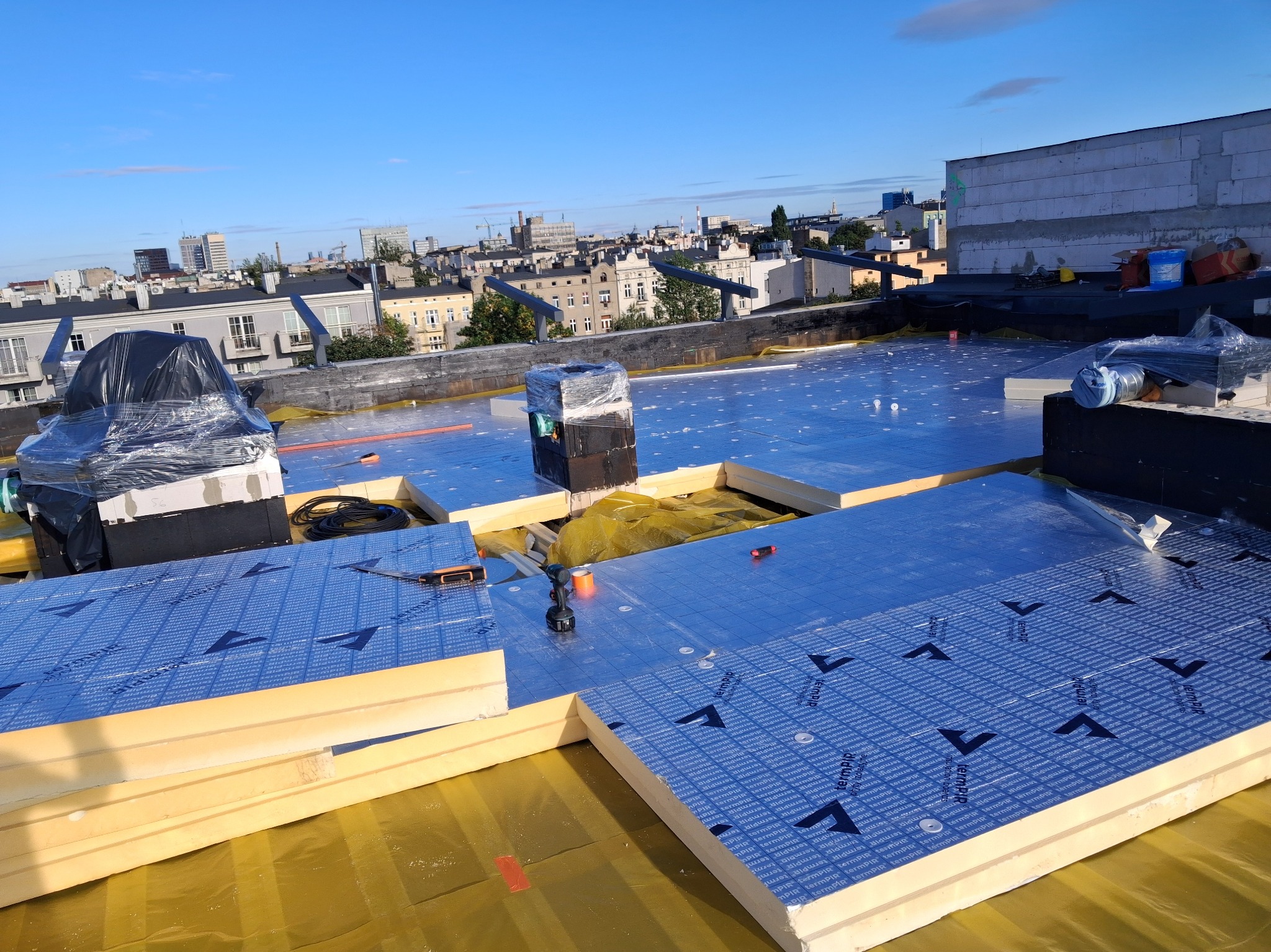 Ocieplenie dachu płaskiego płytami termoizolacyjnymi PIR z folią aluminiową. Widoczne narzędzia i elementy montażowe. W tle zabudowa miejska w słoneczny dzień.