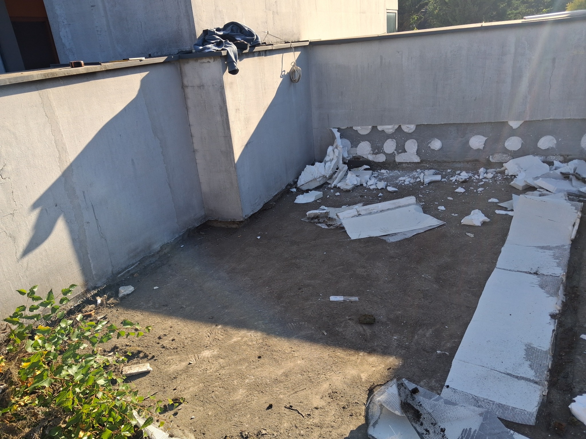 Przygotowanie do izolacji tarasu: fragment ściany z widocznymi otworami, porozrzucane fragmenty styropianu, ziemia. Ujęcie z góry, oświetlenie naturalne.