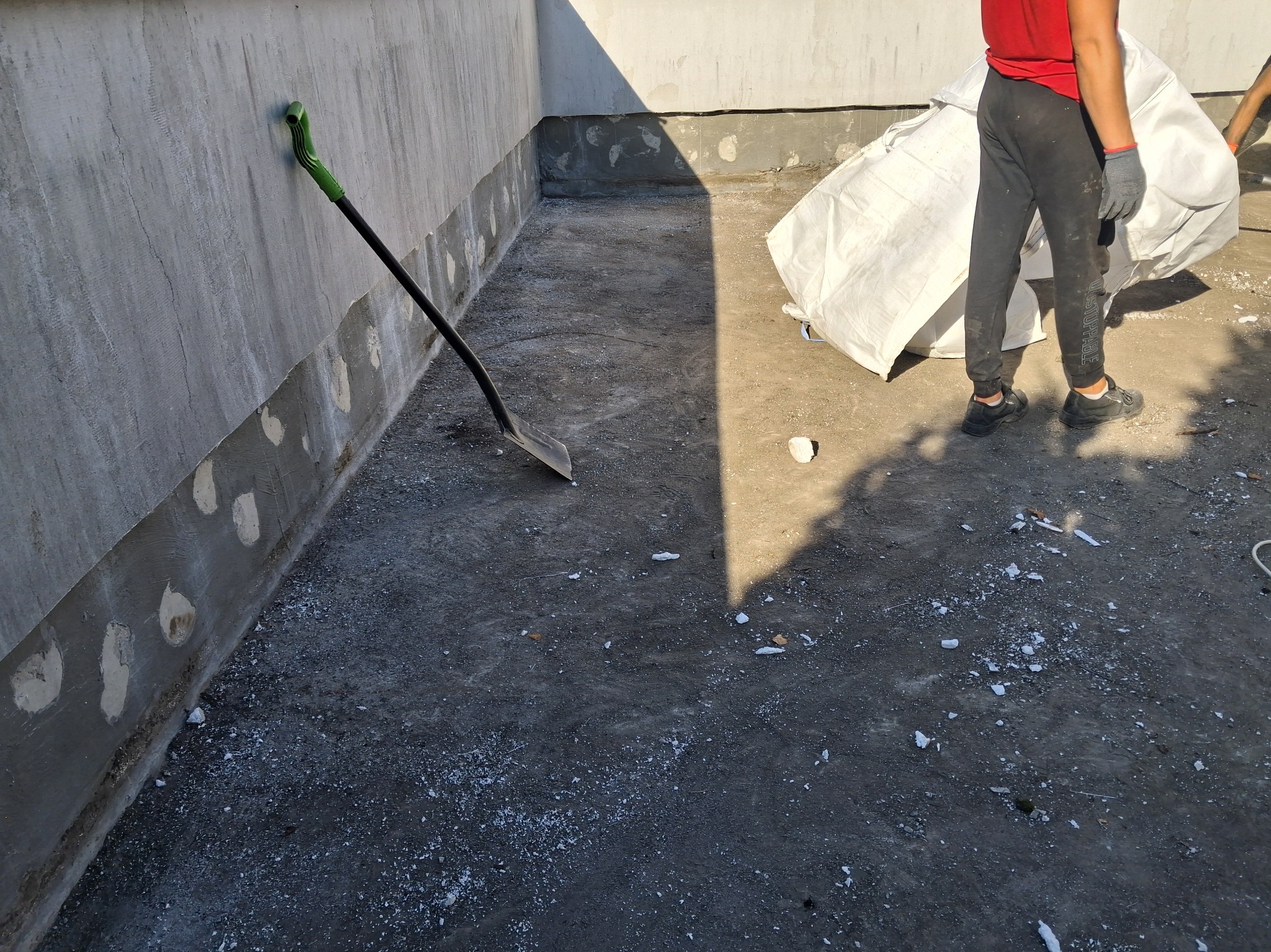 Przygotowanie do izolacji fundamentów: oczyszczona powierzchnia, łopata oparta o ścianę, pracownik z workiem materiału izolacyjnego w tle.