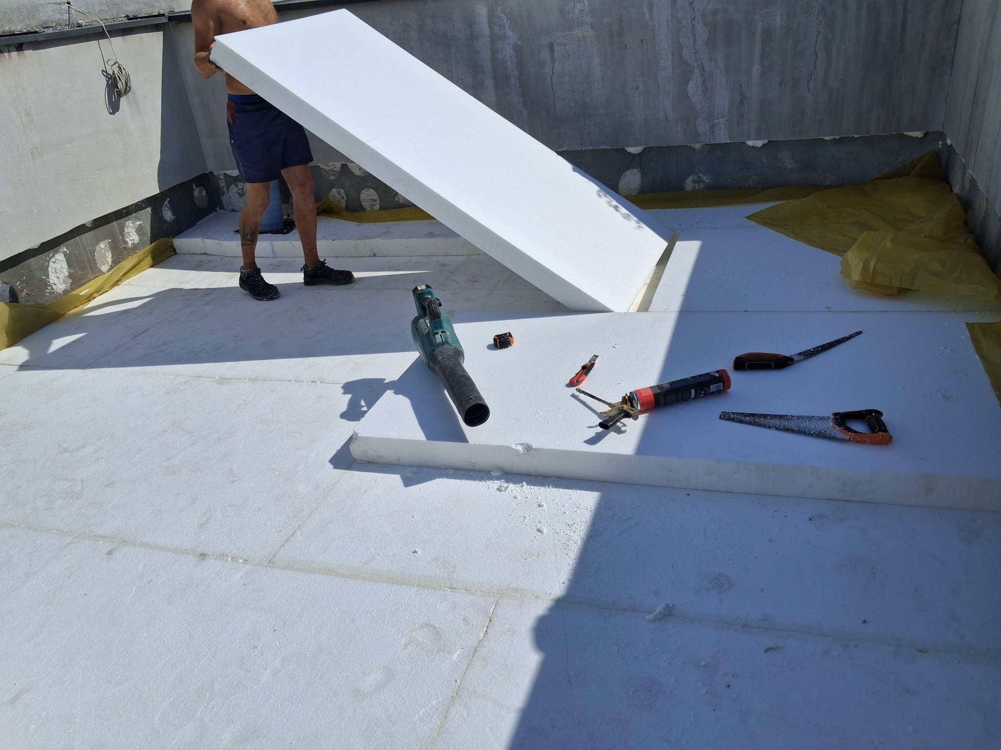 Montaż termoizolacji dachu płaskiego: białe płyty styropianowe układane na żółtej folii, widoczne narzędzia i fragment sylwetki pracownika. Jasne, słoneczne oświetlenie.
