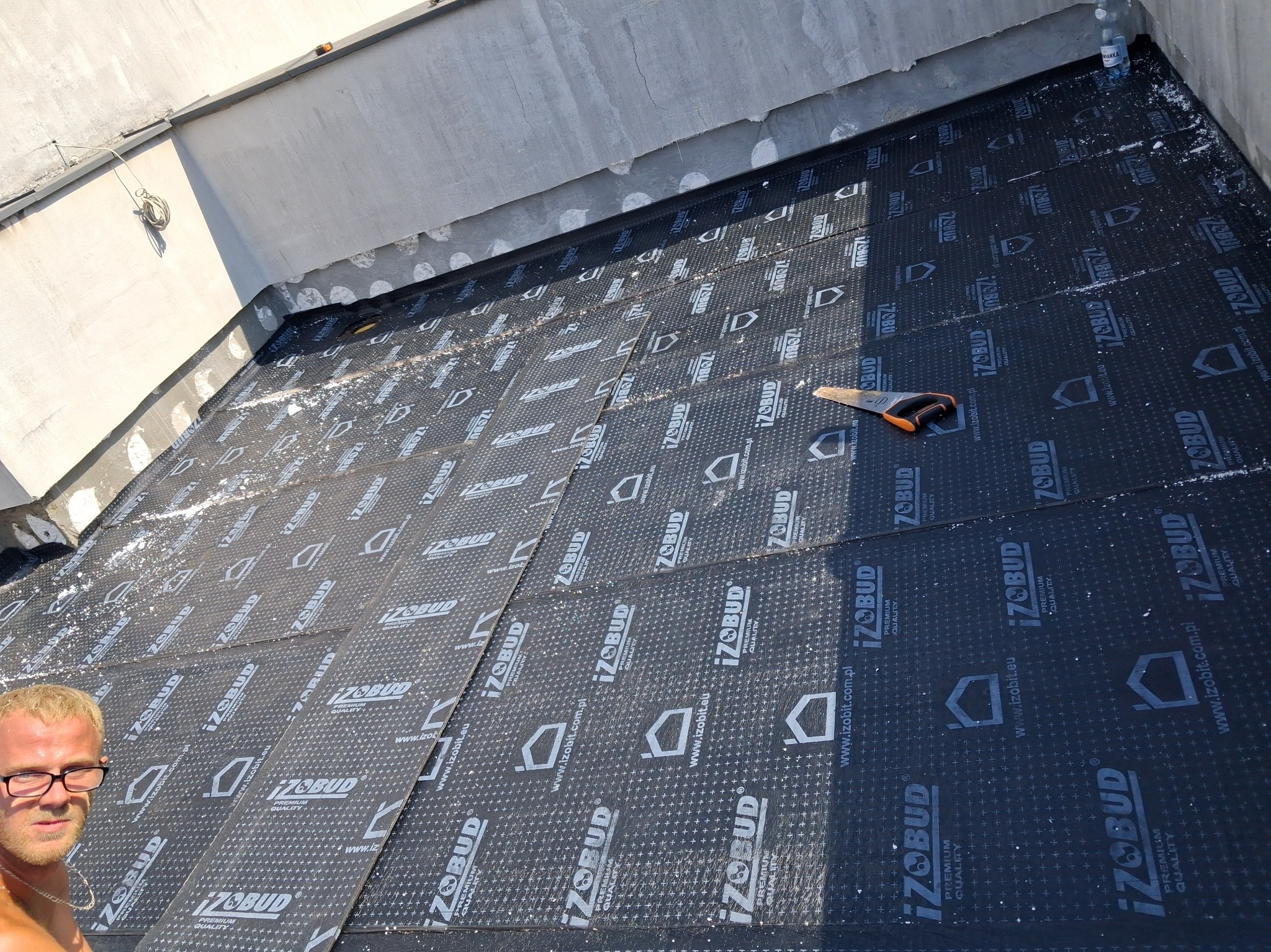 Układanie papy termozgrzewalnej IZOBUD na dachu. Widoczna piła ręczna i fragment osoby. Praca dekarska w trakcie, z bliska, z detalami materiału.