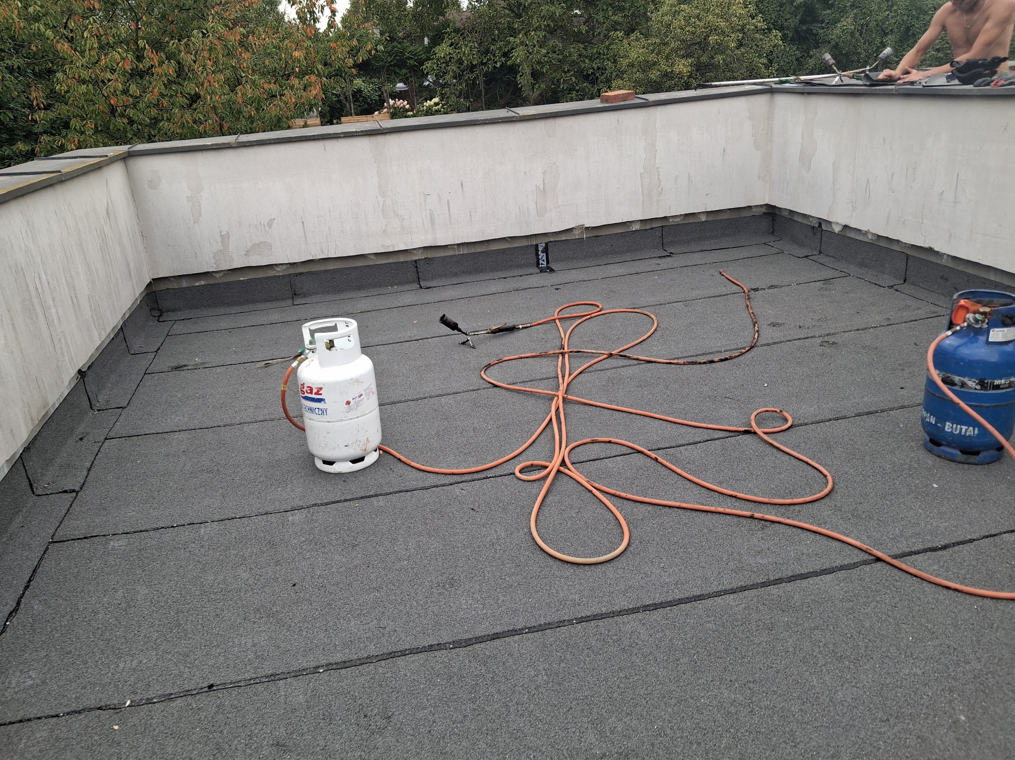 Prace dekarskie na płaskim dachu: biała butla gazowa, palnik dekarski z pomarańczowym wężem. W tle fragment budynku i zieleń drzew. Widoczny dekarz przy pracy.