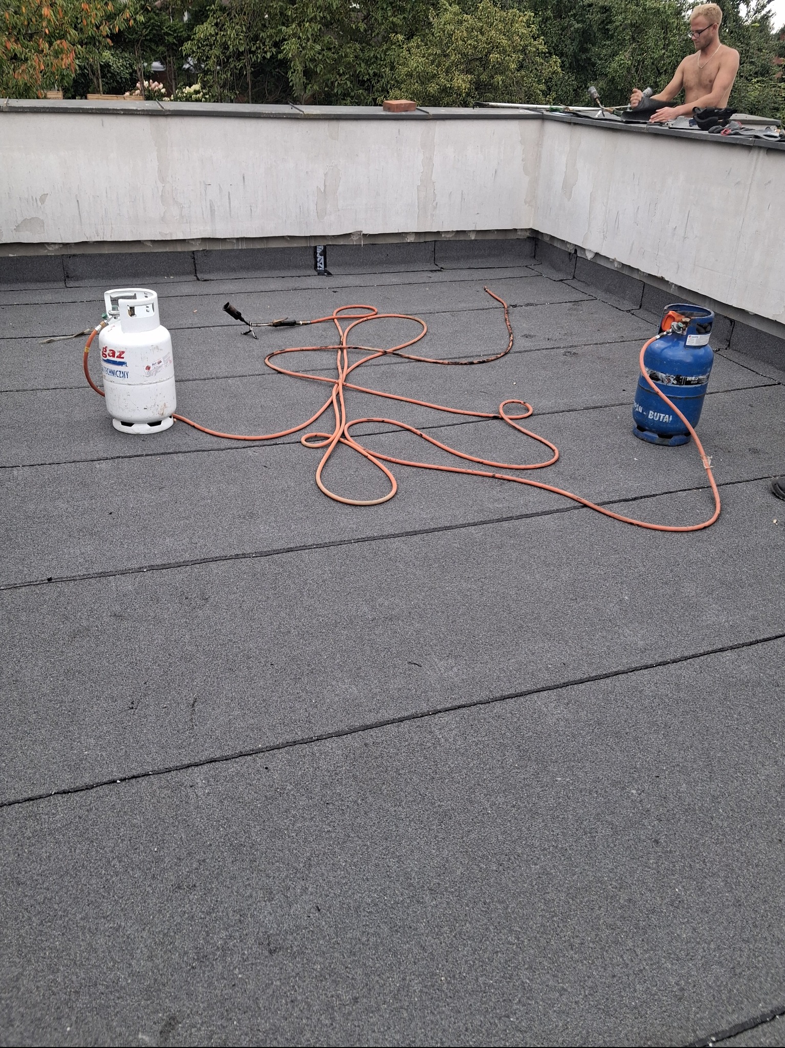 Praca na dachu płaskim: dekarz z palnikiem i butlami gazowymi na tle zieleni. Widoczna papa dachowa i narzędzia. Ujęcie z góry, podkreślające detale wykonania.