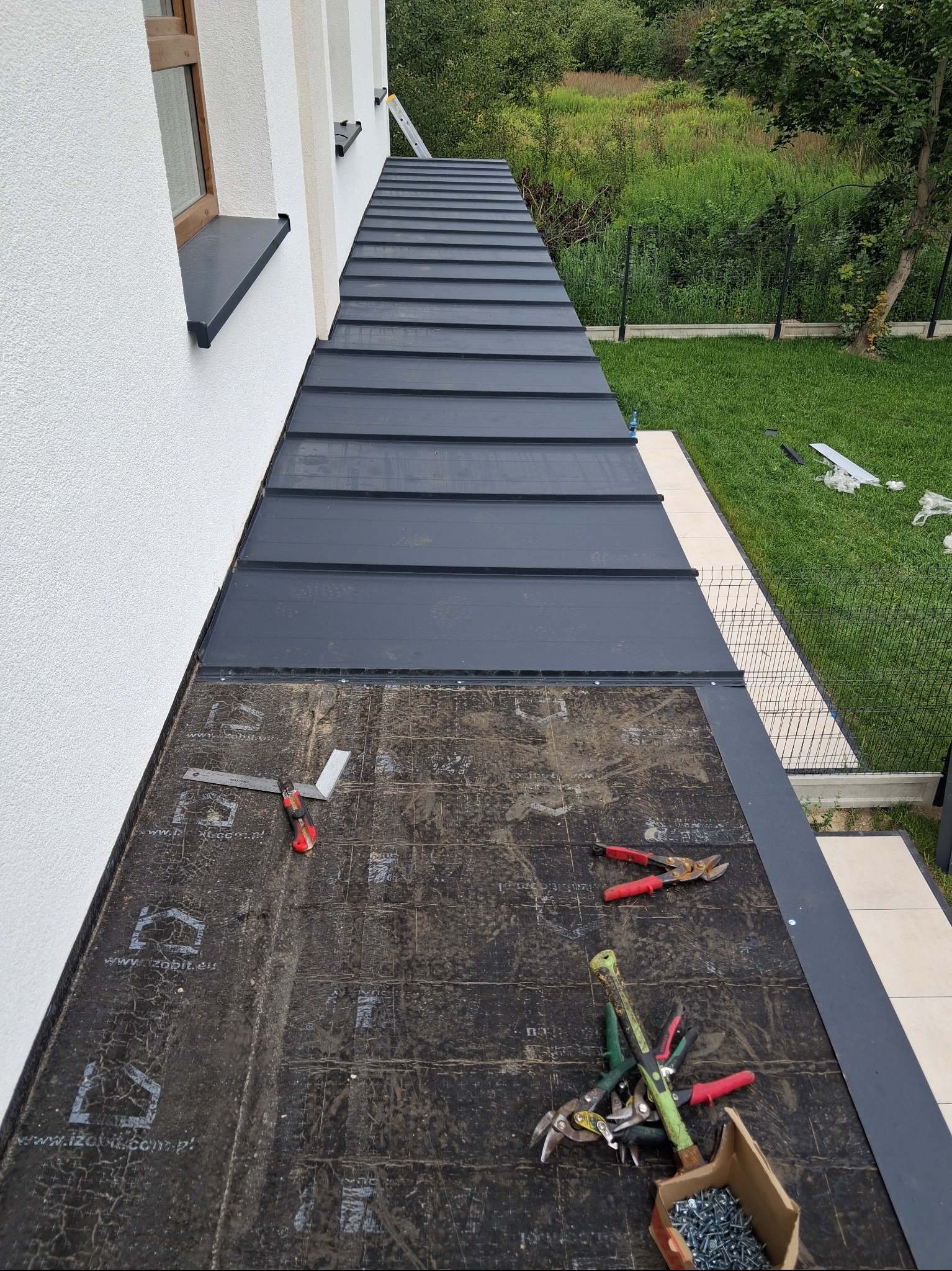 Montaż blachy na dachu domu jednorodzinnego. Widoczne narzędzia dekarskie, fragment pokrycia i podkład. Ujęcie z góry, z bliska, w dziennym świetle.