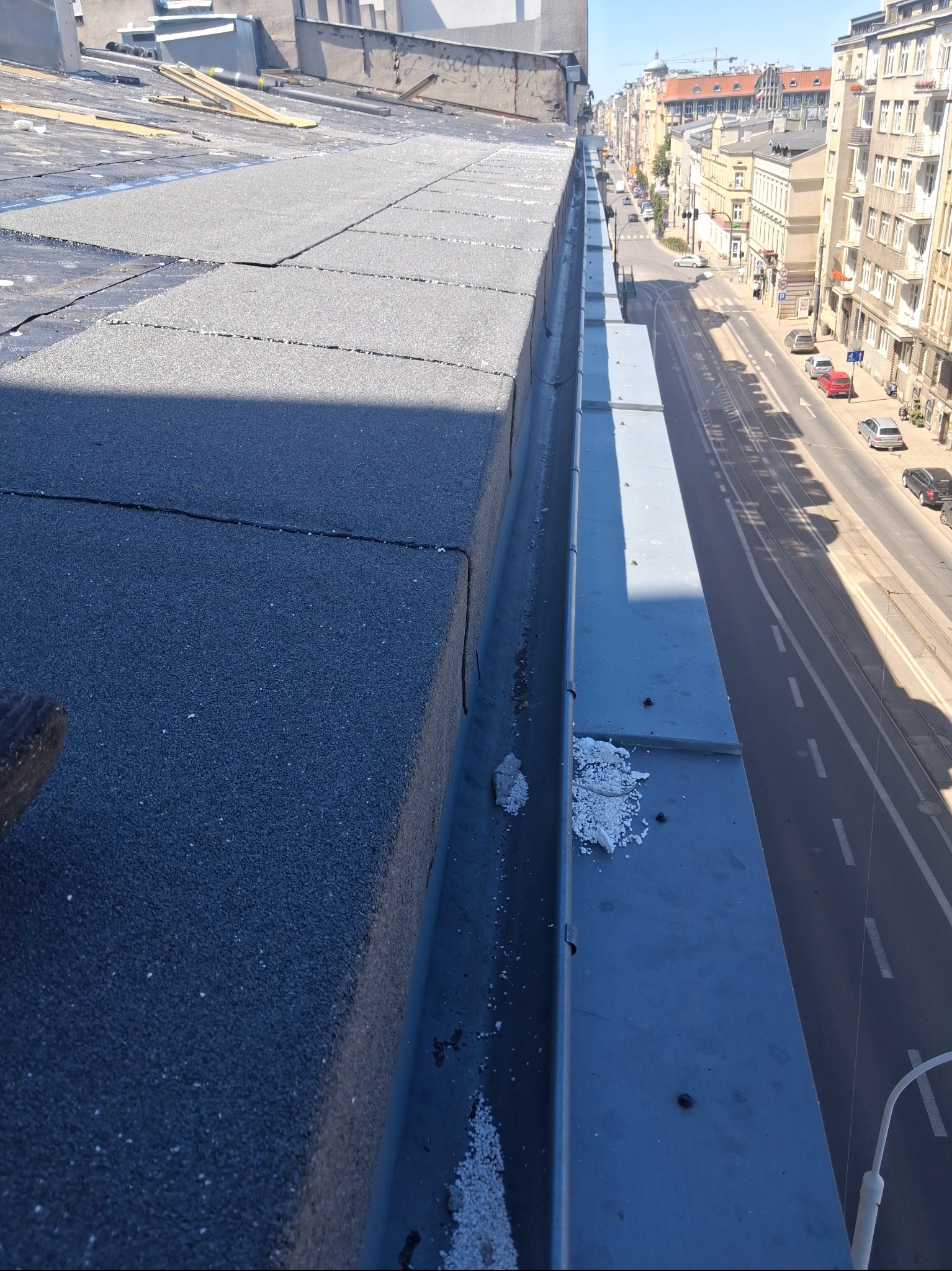 Fragment dachu budynku z widoczną nową, szarą papą bitumiczną i rynną. W tle ulica miejska z budynkami w słoneczny dzień. Widok z góry.