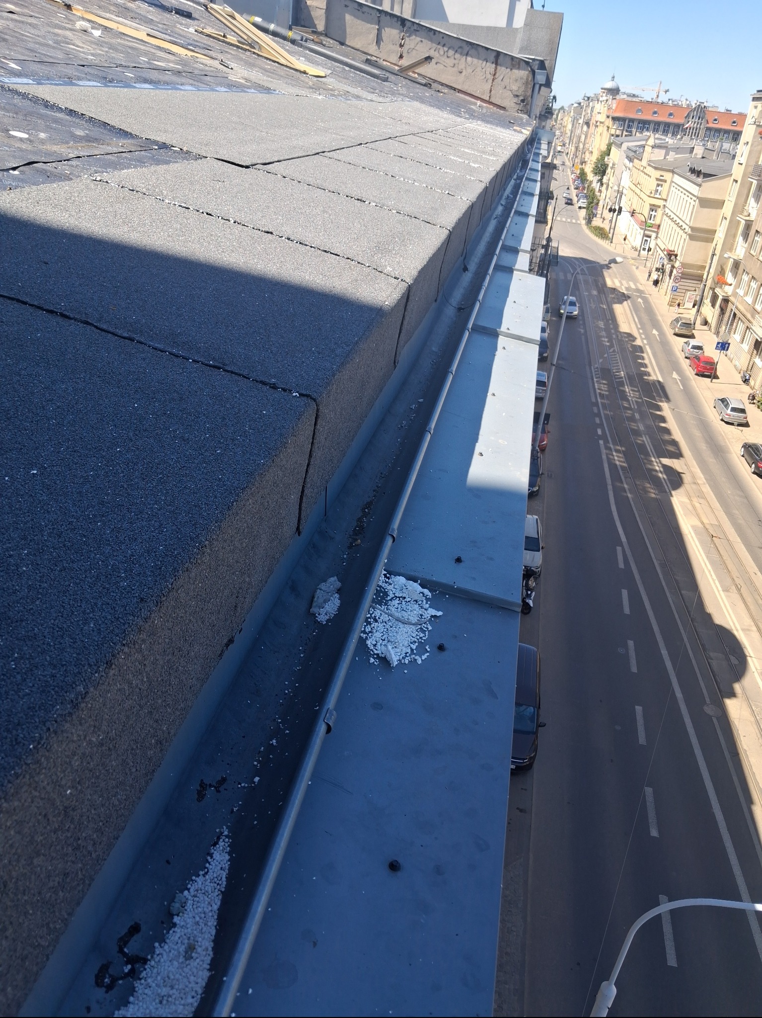 Widok z góry na fragment dachu pokrytego papą i rynnę z zanieczyszczeniami. W tle zabudowa miejska i ulica z samochodami. Słoneczny dzień.