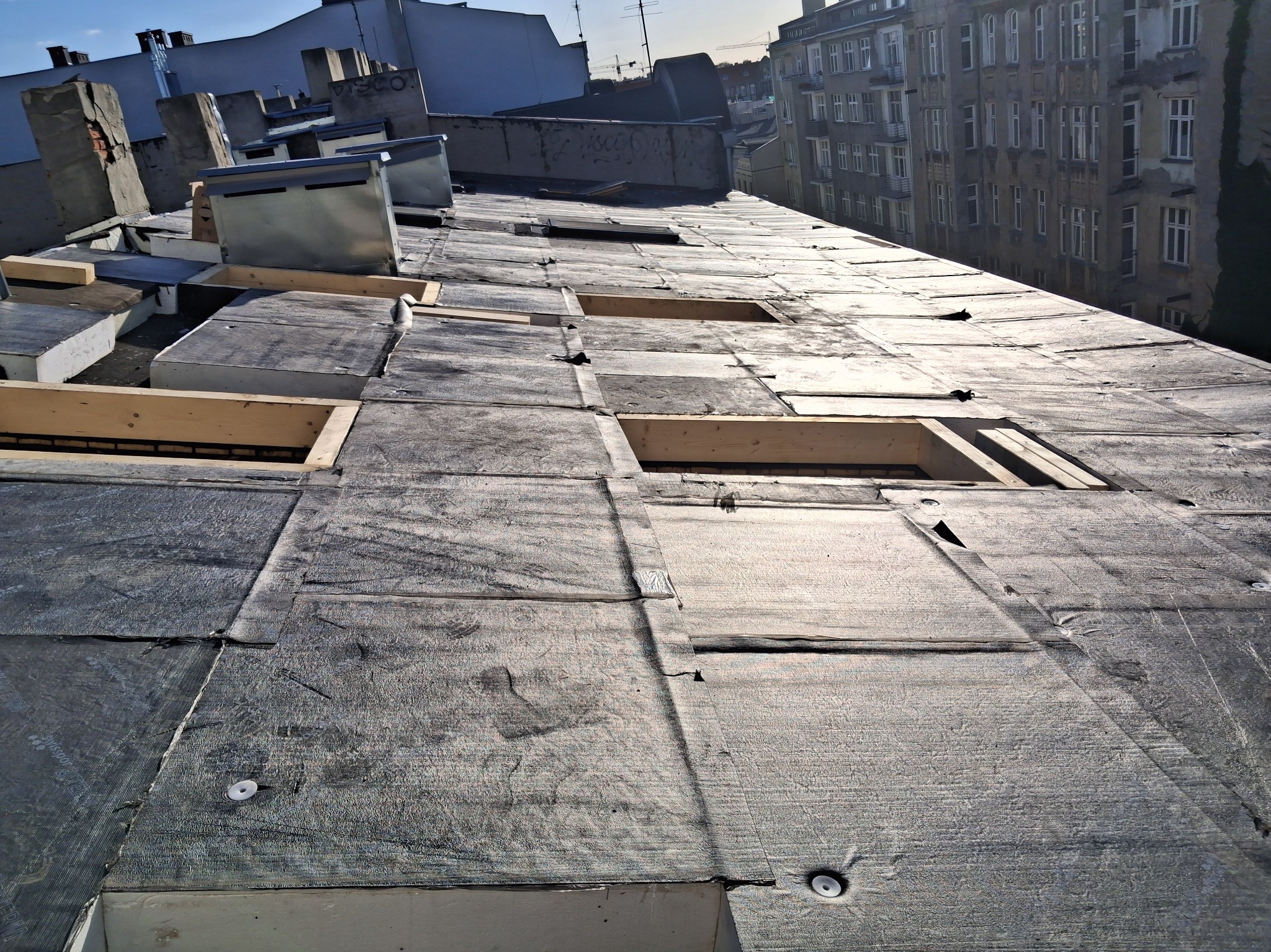 Dach pokryty papą termozgrzewalną z widocznymi łatami i elementami drewnianej konstrukcji. W tle budynki miejskie w słoneczny dzień.
