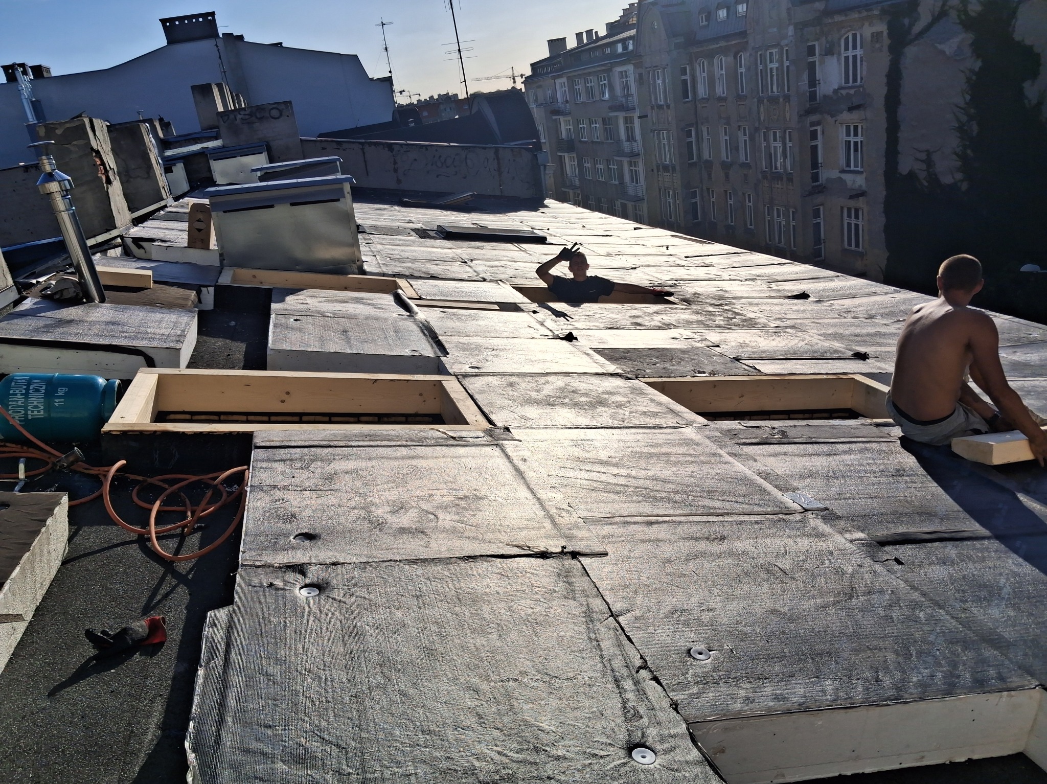 Prace dekarskie na dachu kamienicy: montaż izolacji termicznej z widocznym pracownikiem w otworze i butlą gazową. Detaliczne ujęcie na etapie ocieplania.