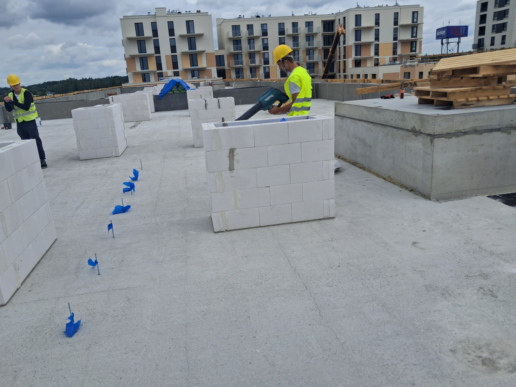 Pracownik w żółtym kasku i kamizelce łączy bloczki betonu komórkowego na dachu budynku wielorodzinnego przy użyciu specjalistycznego narzędzia.