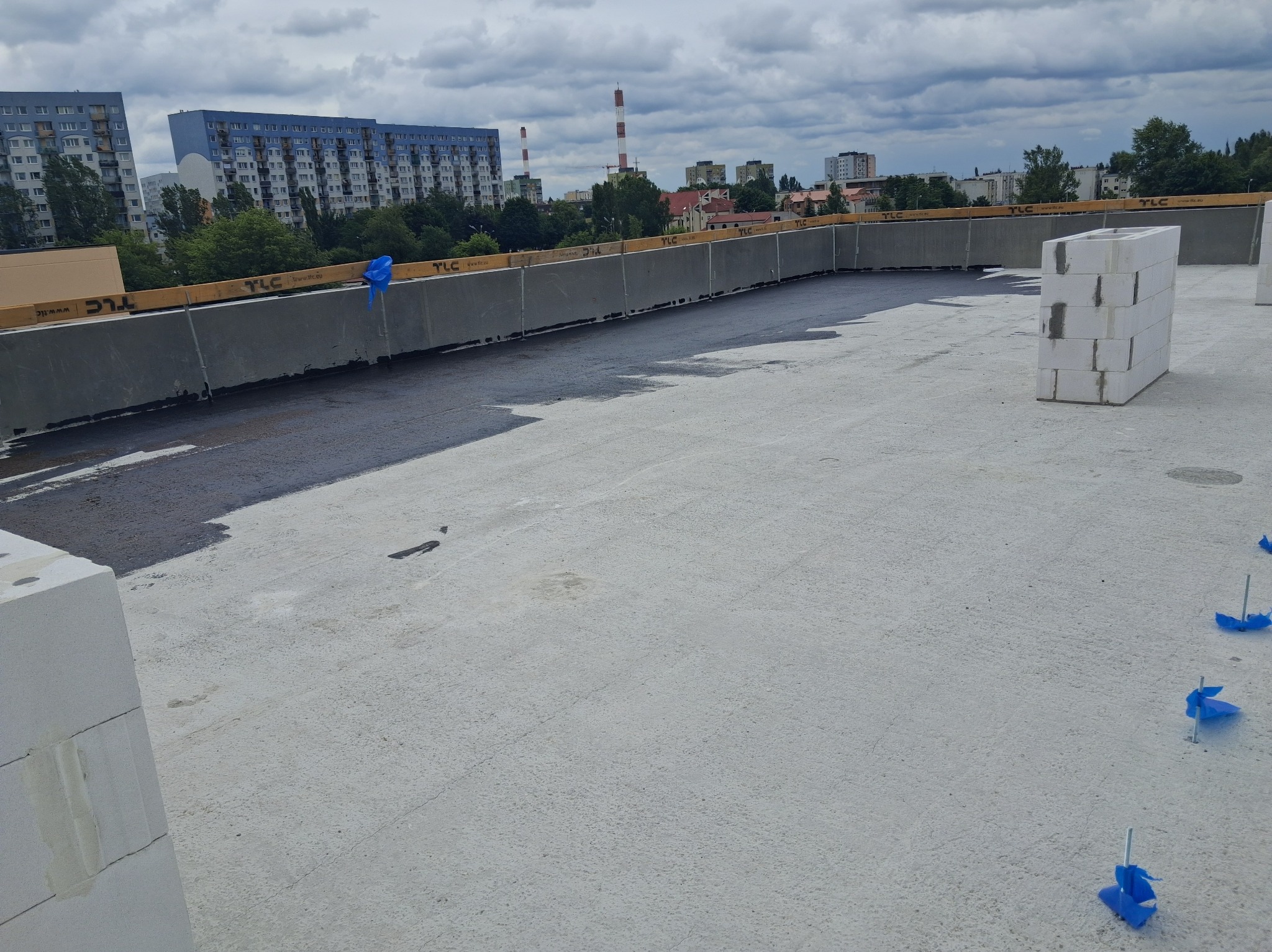 Płaski dach pokryty częściowo czarną izolacją, z widocznymi bloczkami Ytong i elementami mocującymi. W tle budynki mieszkalne i pochmurne niebo.