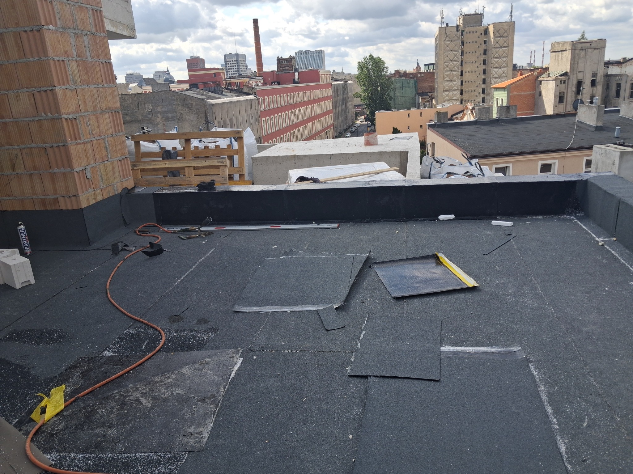 Fragment dachu z papy termozgrzewalnej w trakcie prac dekarskich. Widoczne narzędzia, elementy papy i panorama miasta w tle. Ujęcie z góry.