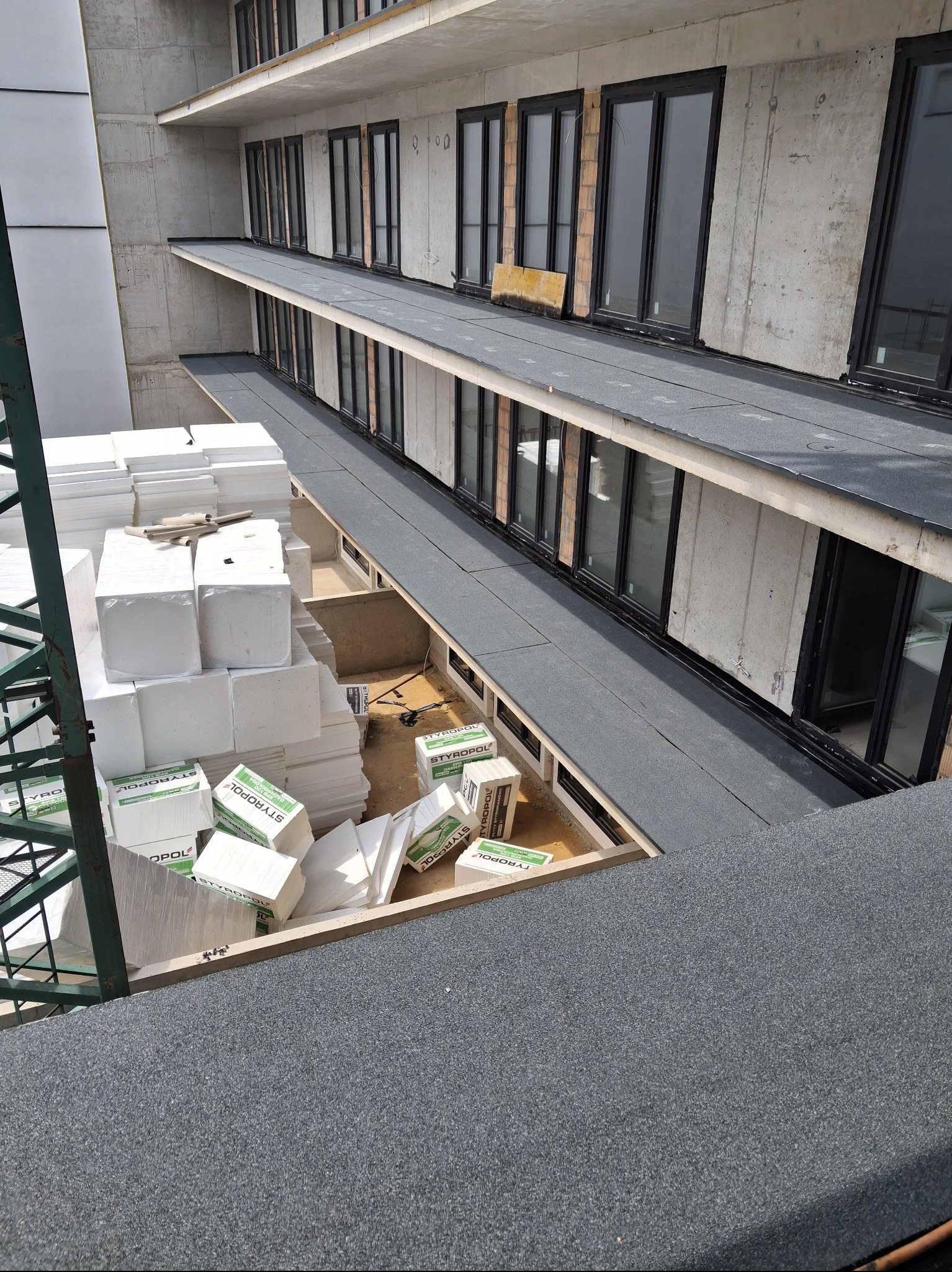 Budowa: izolacja termiczna dachu budynku wielorodzinnego. Styropian w opakowaniach i rozpakowany, widok z góry na płaski dach z oknami. Etap prac izolacyjnych.