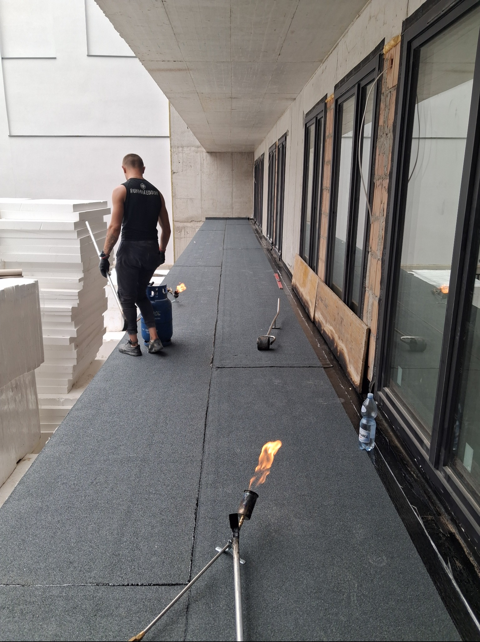 Pracownik w trakcie hydroizolacji dachu płaskiego papą termozgrzewalną. Widoczne narzędzia, butla gazowa i zapas styropianu. Dach w surowym stanie.