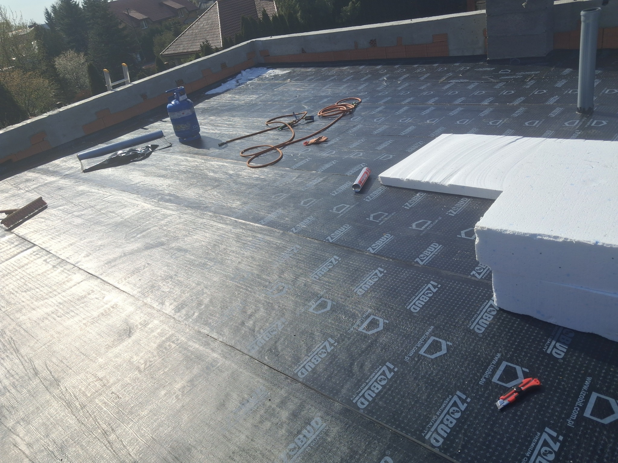 Prace dekarskie: membrana hydroizolacyjna z widocznym nadrukiem, butla gazowa, narzędzia dekarskie i fragmenty styropianu na płaskim dachu w trakcie realizacji.