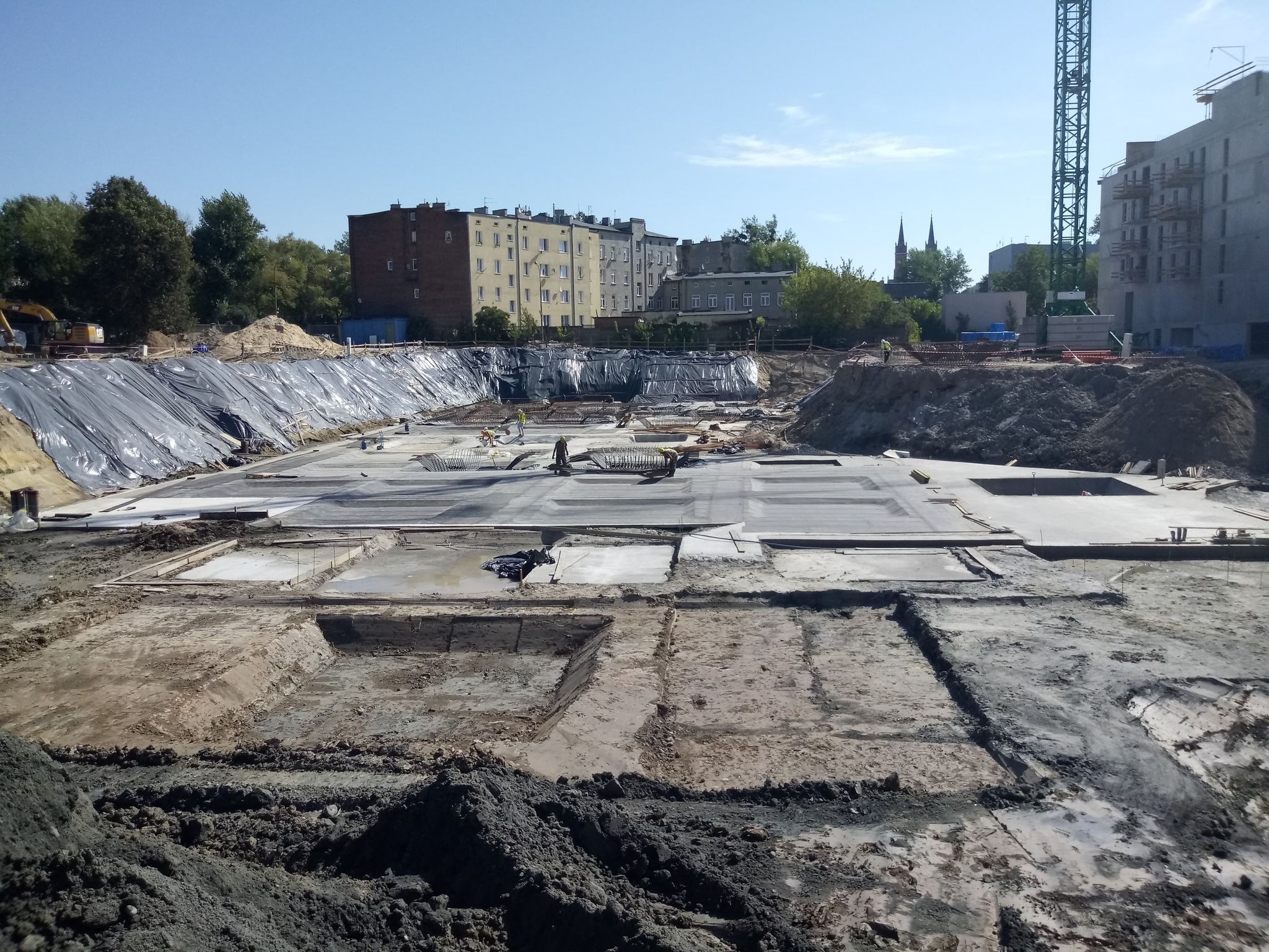 Budowa fundamentów budynku w otwartym wykopie, widoczne zbrojenia, betonowe płyty i czarna folia zabezpieczająca, w tle budynki mieszkalne i żuraw budowlany.