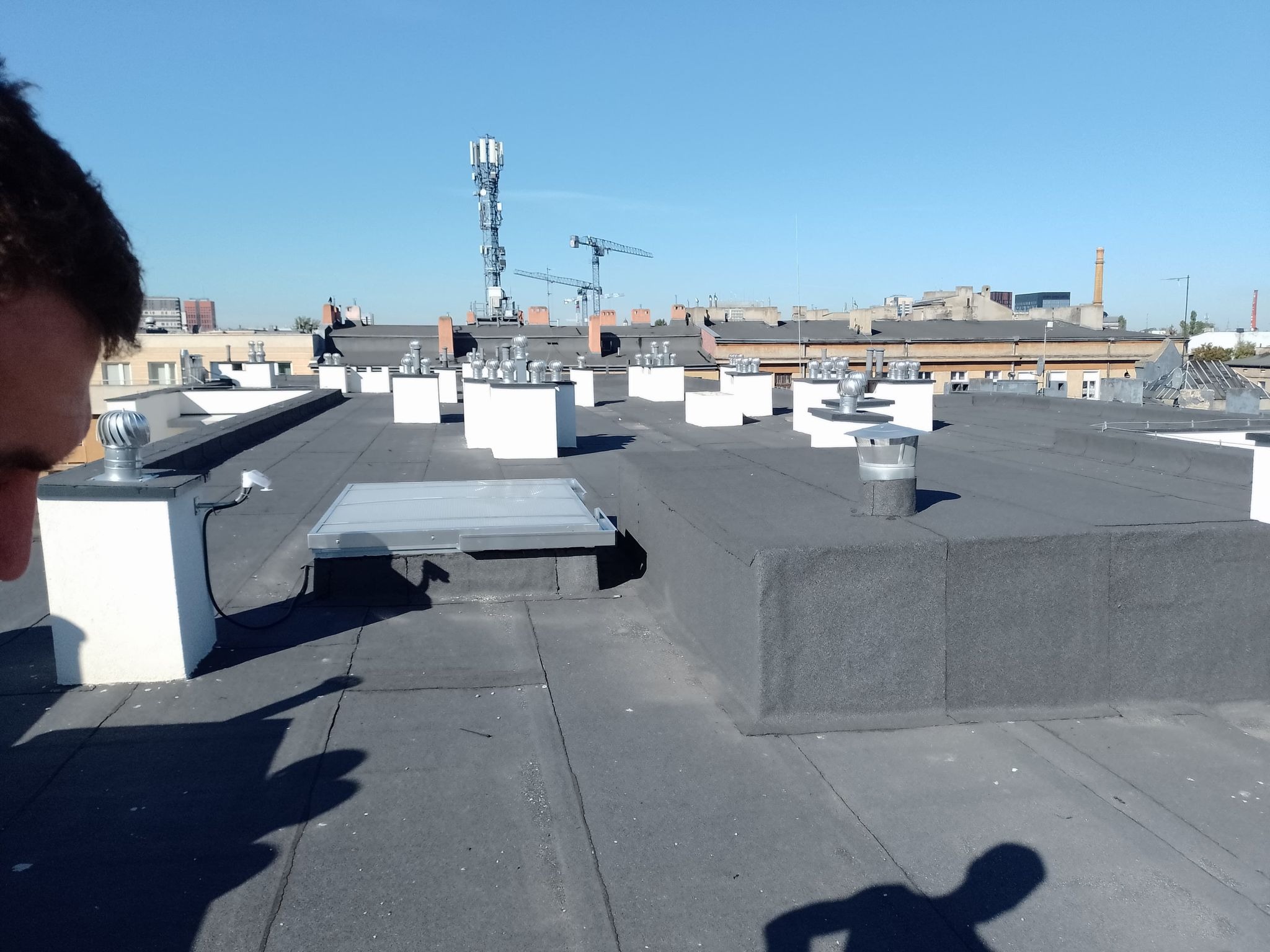 Płaski dach pokryty papą termozgrzewalną z licznymi kominami wentylacyjnymi i wyłazem dachowym na tle miejskiego krajobrazu z żurawiami budowlanymi i wieżą telekomunikacyjną.