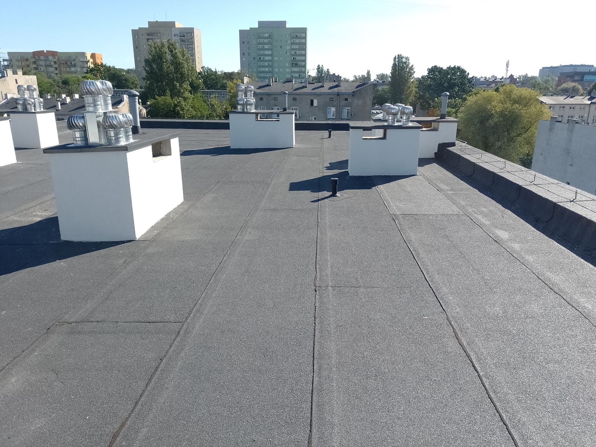 Płaski dach pokryty papą termozgrzewalną z kominami wentylacyjnymi, budynki w tle, widok z góry.