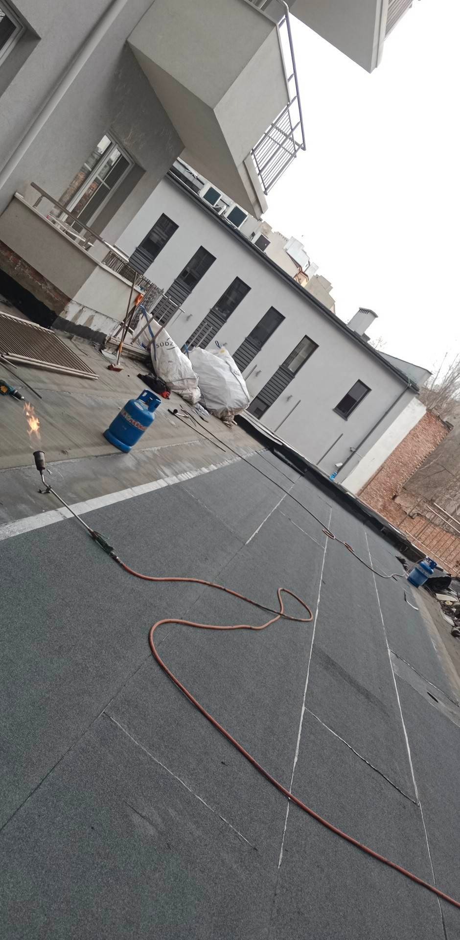 Prace dekarskie na dachu budynku wielorodzinnego, widoczny palnik dekarski, butla gazowa, zgrzewana papa na dachu, białe worki z odpadami budowlanymi w tle.