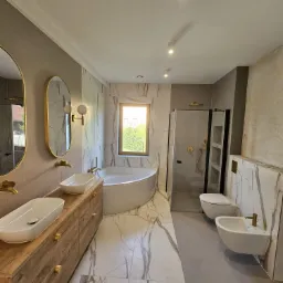 Nowoczesna łazienka z marmurowymi płytkami, drewnianą szafką pod umywalki z owalnymi umywalkami nablatowymi, złotą armaturą, owalnymi lustrami w złotych ramach, wanną przy oknie i kabiną...