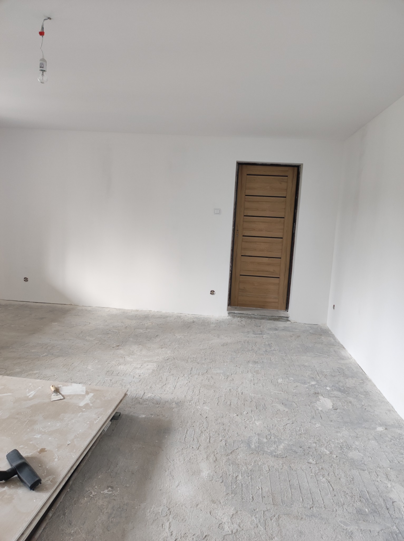 Pokój w trakcie remontu, z drewnianymi drzwiami, surową podłogą po wylewce, nieotynkowanymi ścianami, wiszącą żarówką i stertą płyt gipsowych z narzędziami w rogu.