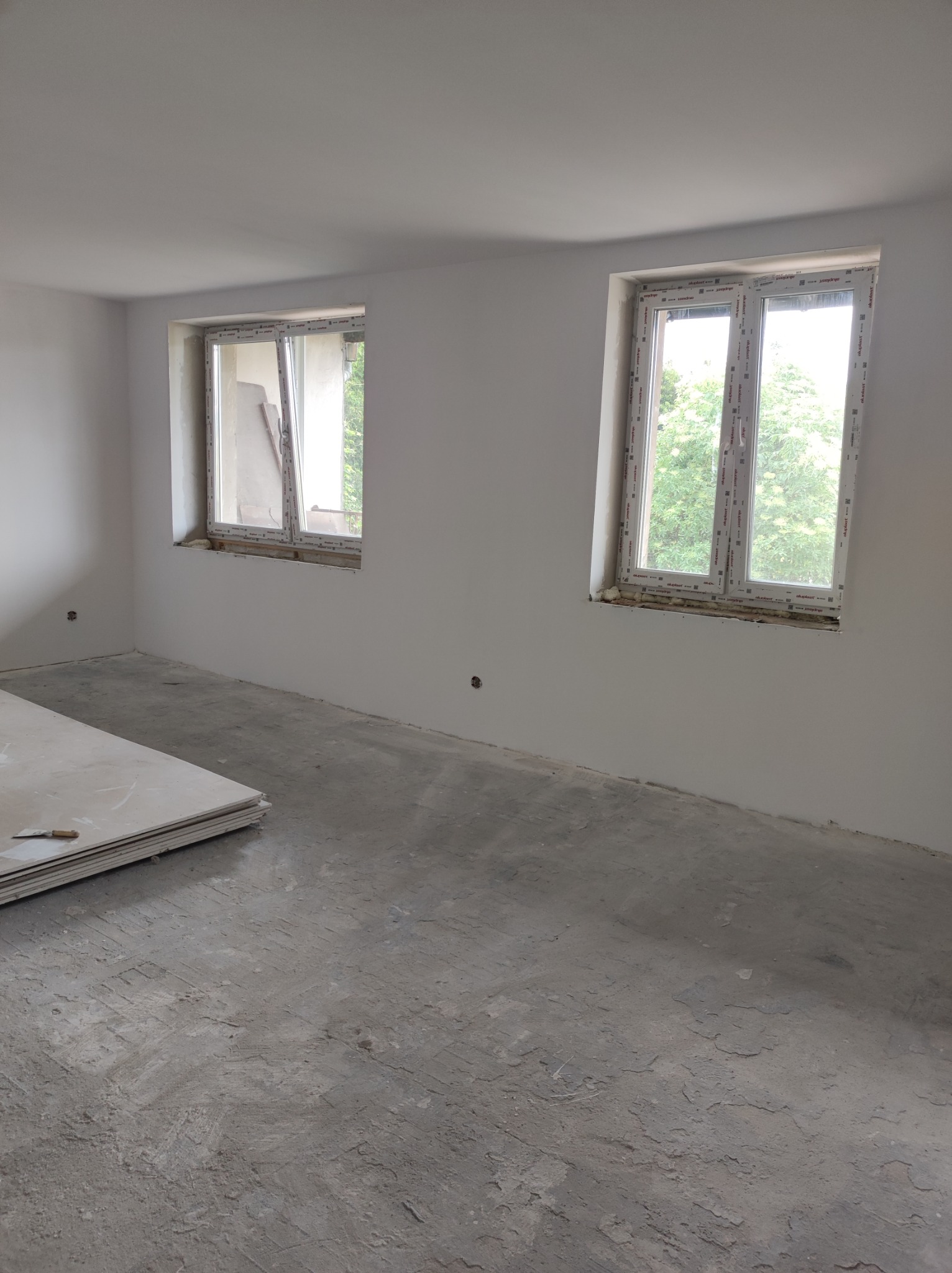 Pokój w trakcie remontu z dwoma oknami PCV, jedno z nich uchylone, na podłodze stos płyt karton-gips z widoczną szpachelką.