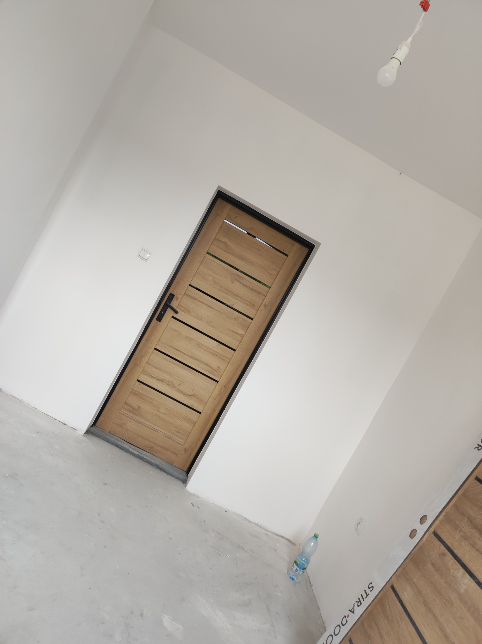 Nowo zamontowane drzwi wewnętrzne z poziomymi, ciemnymi wstawkami na tle świeżo pomalowanych, białych ścian w trakcie remontu, widoczna butelka wody na podłodze i prowizoryczne oświetlenie.