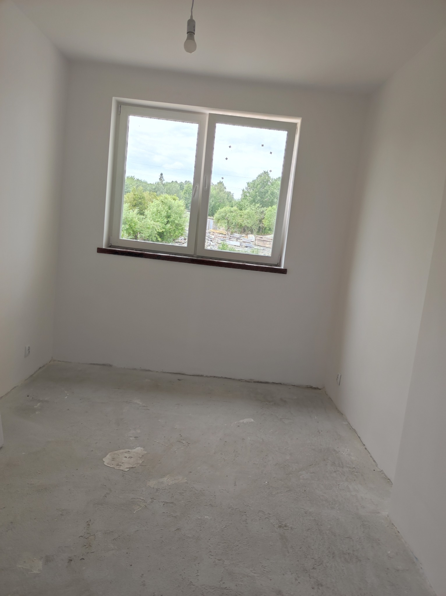 Pusty, jasny pokój z białymi ścianami i surowym betonowym podłożem, okno z widokiem na zieleń, pojedyncza żarówka zwisająca z sufitu.