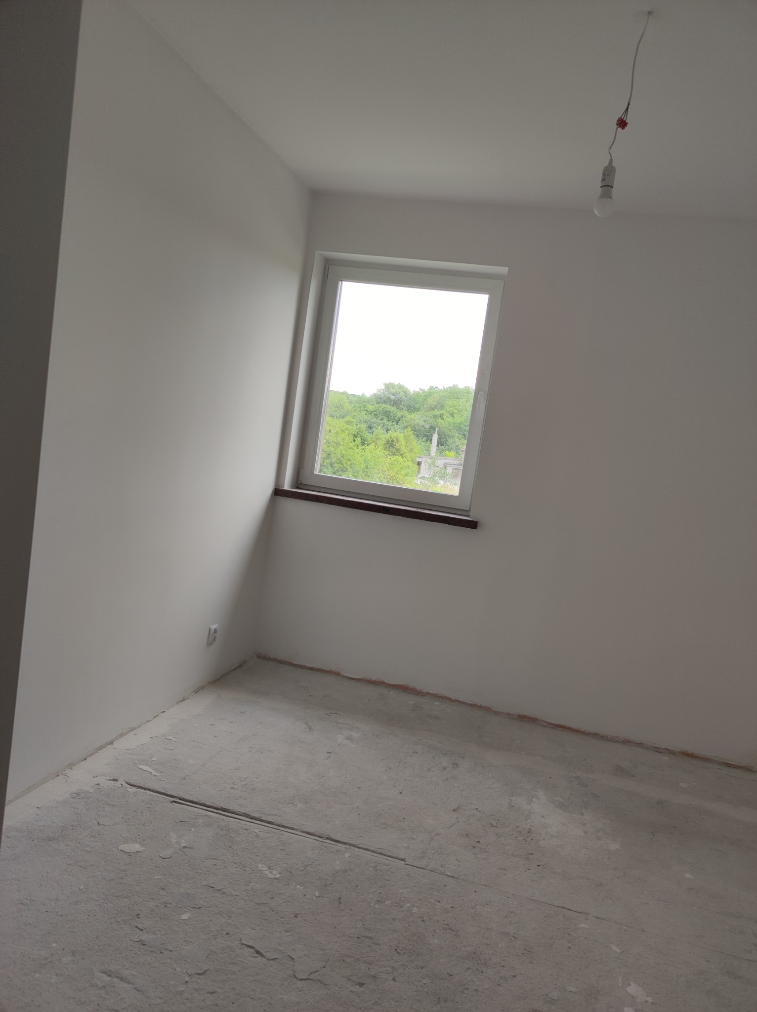 Surowe wnętrze pokoju z oknem, ściany świeżo pomalowane na biało, widoczny surowy beton na podłodze oraz wystające przewody z żarówką.