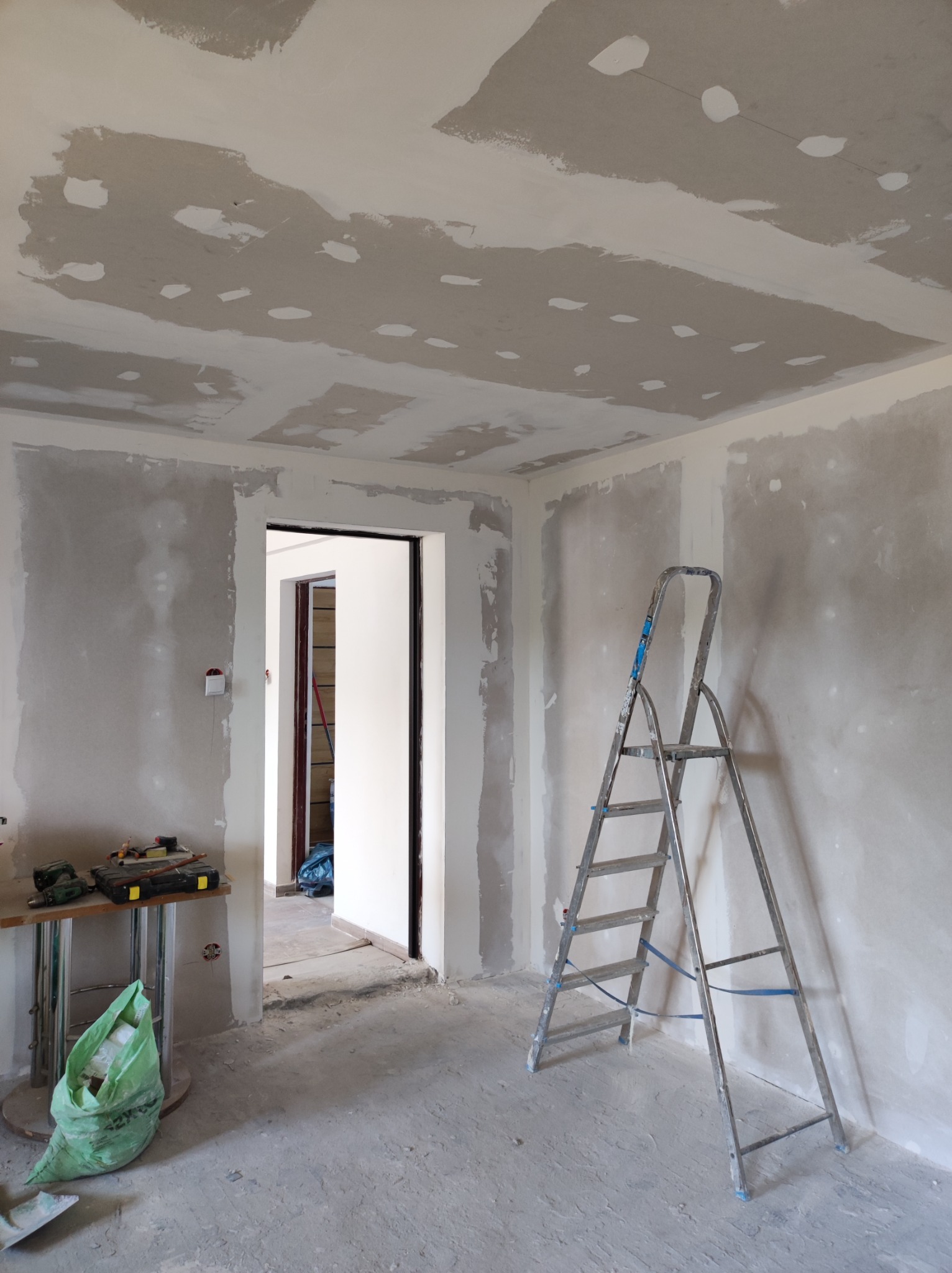 Pokój w trakcie wykańczania: ściany i sufit pokryte warstwą szpachli, widoczna drabina aluminiowa, stół z narzędziami i workiem na śmieci, w tle otwarte drzwi do kolejnego pomieszczenia.