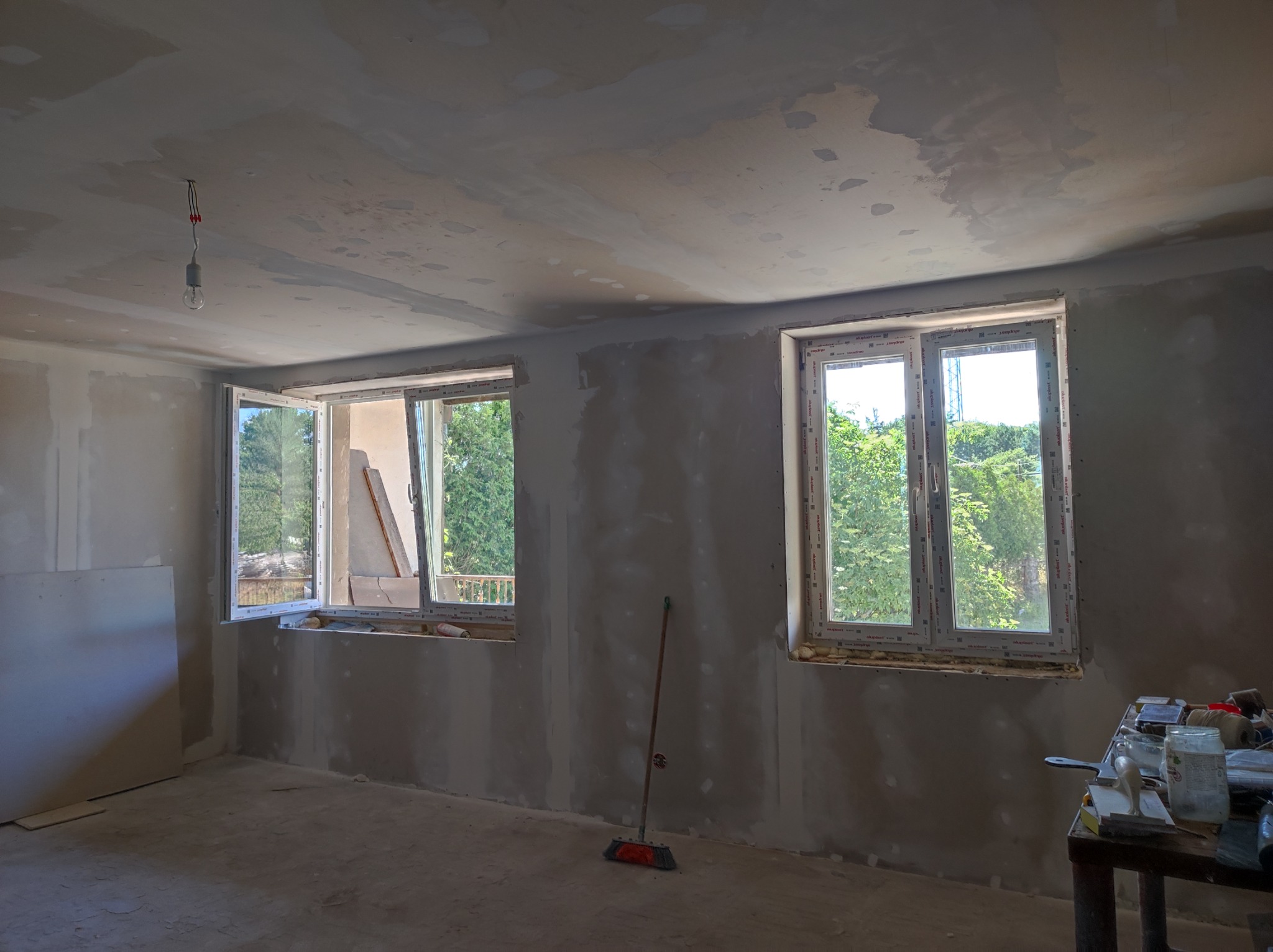 Pokój w trakcie remontu: ściany i sufit po nałożeniu gładzi, dwa okna z widokiem na zieleń, jedno z nich uchylone, na podłodze szczotka, na stole narzędzia i materiały budowlane.