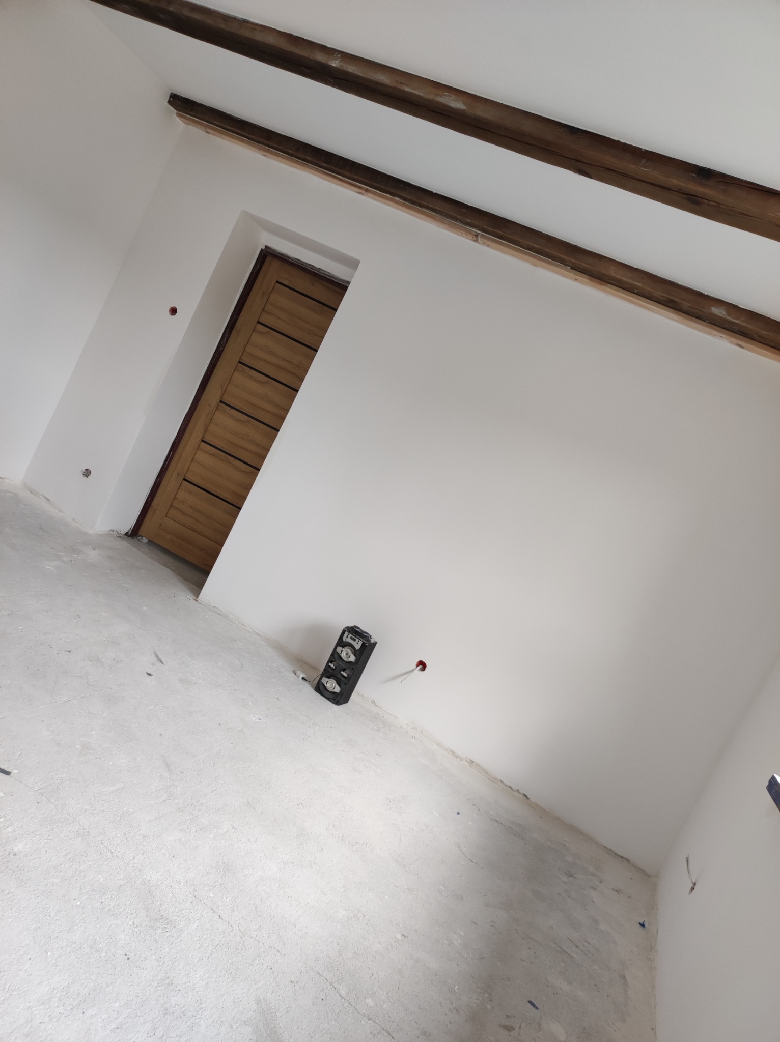 Wnętrze w trakcie remontu: białe ściany z drewnianymi belkami sufitowymi, drewniane drzwi z poziomymi panelami, poziomica na podłodze, ślady po pracach wykończeniowych.