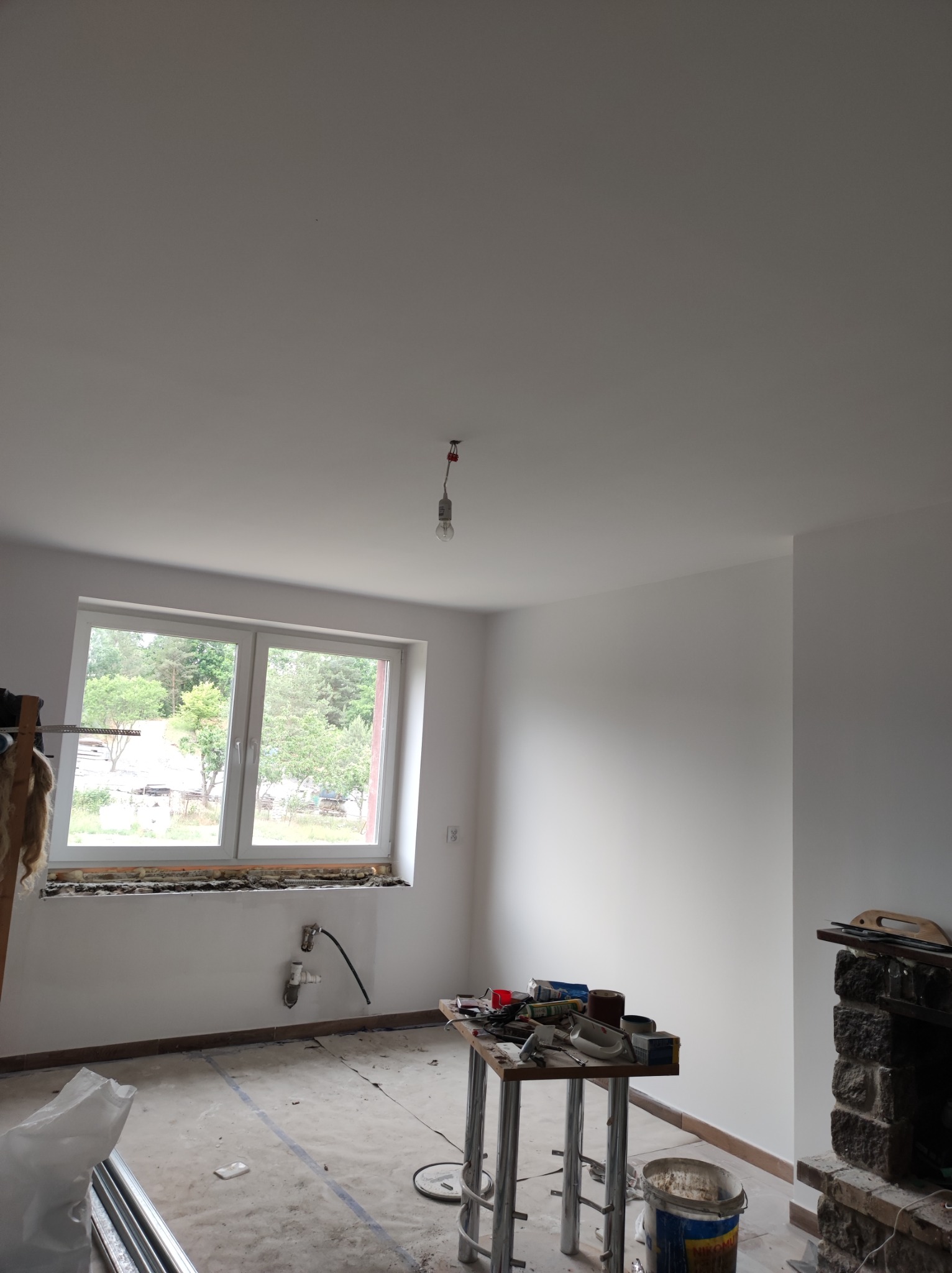 Wnętrze pokoju w trakcie remontu, z widocznym oknem po wymianie, narzędziami na stole roboczym, oraz przygotowanym do prac wykończeniowych kominkiem z kamienia.