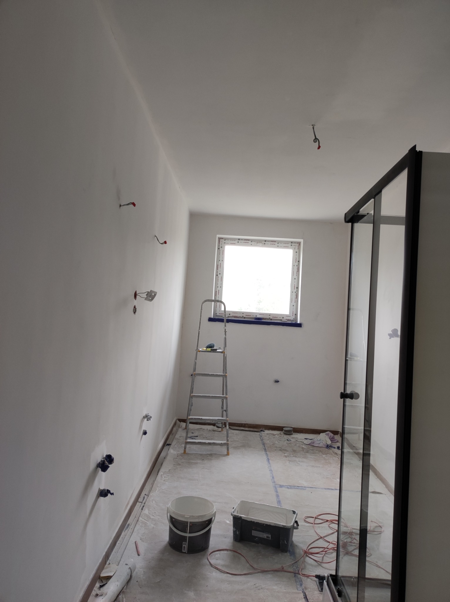 Wnętrze łazienki w trakcie malowania na biało, widoczne wystające kable, drabina malarska, wiadro z farbą i kuweta malarska, oraz częściowo widoczna kabina prysznicowa z czarnymi ramami.