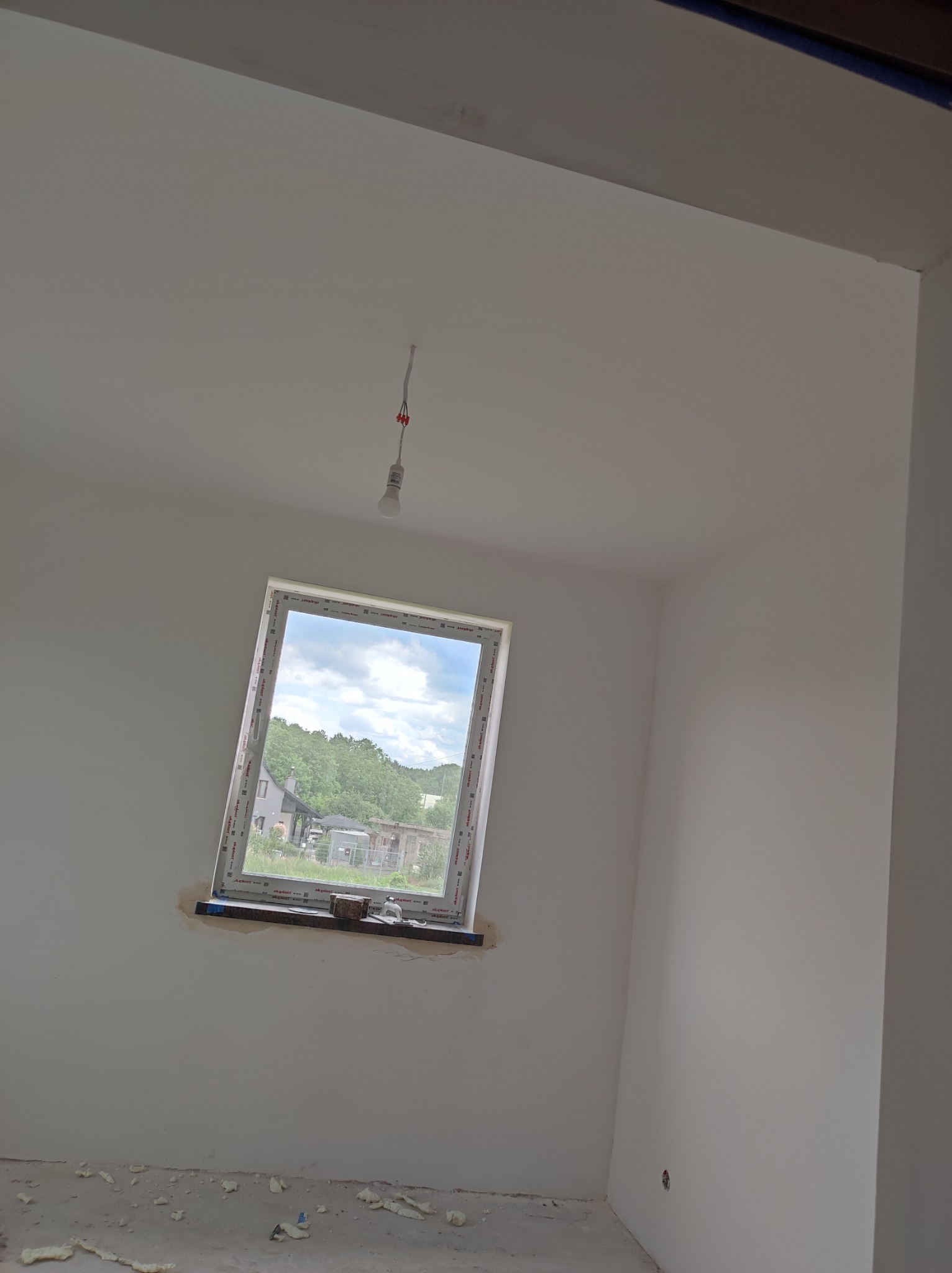 Wnętrze pokoju po remoncie, świeżo pomalowane ściany na biało, zamontowane okno z widokiem na zieleń, widoczne ślady prac na podłodze i wokół okna, surowa żarówka zwisająca z sufitu.