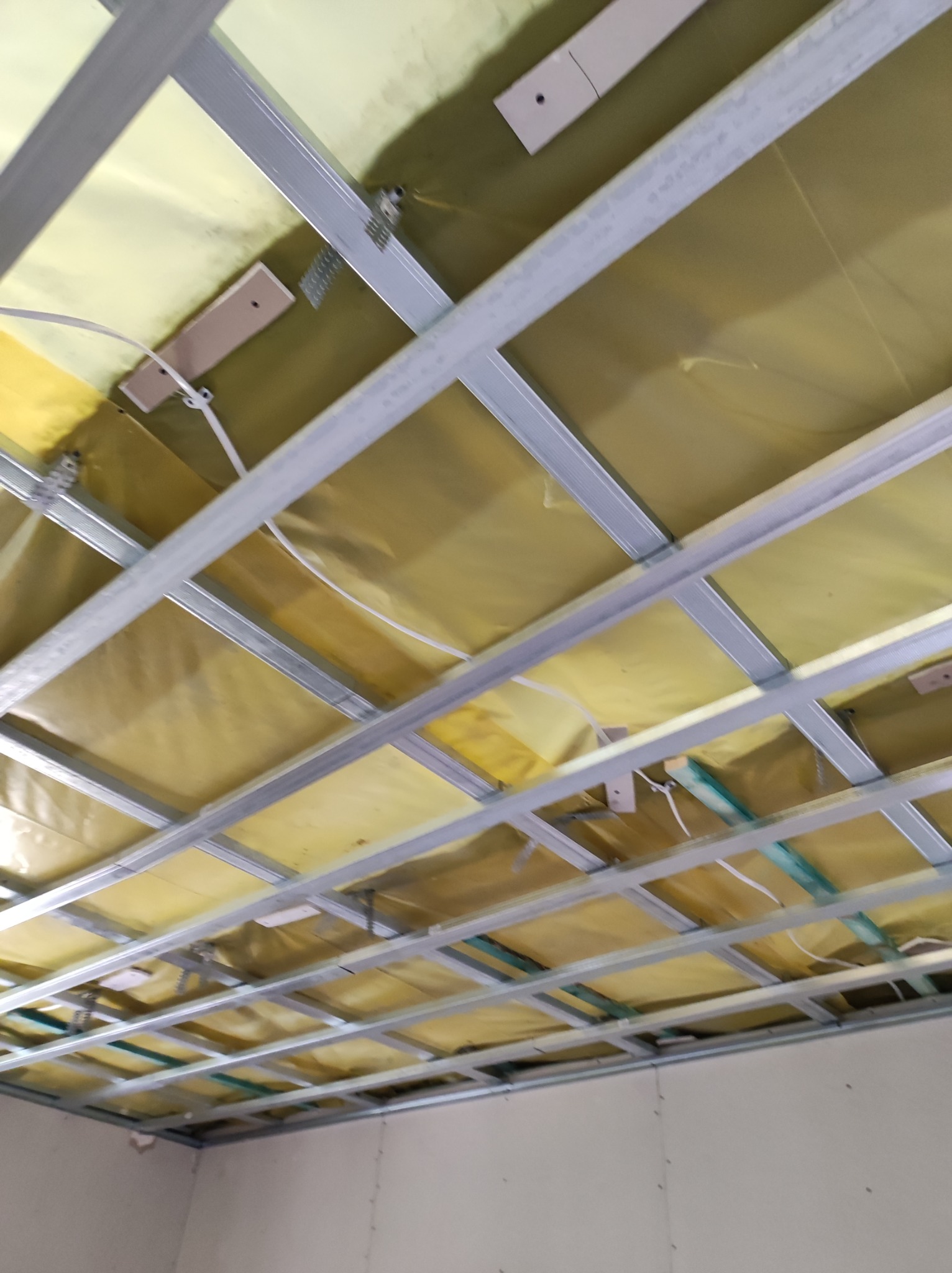 Stelaż sufitowy z profili metalowych, wypełniony żółtą izolacją, widok z dołu w trakcie montażu, przewody elektryczne widoczne.