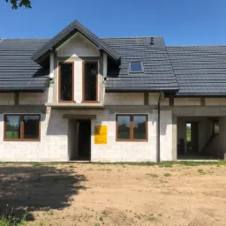 Surowy stan budynku jednorodzinnego z szarej dachówki i brązowymi ramami okiennymi, widoczne pustaki i plac budowy.