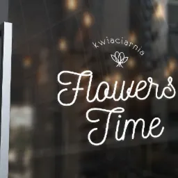 Biały napis 'kwiaciarnia Flowers Time' na szklanych drzwiach wejściowych, widoczne srebrne uchwyty i odbicie wnętrza.