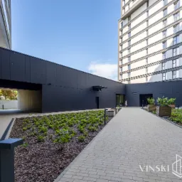 Dziedziniec nowoczesnego apartamentowca z geometryczną architekturą, wyłożony jasną kostką brukową, z rabatami obsadzonymi niską zielenią i drzewami w drewnianych donicach.