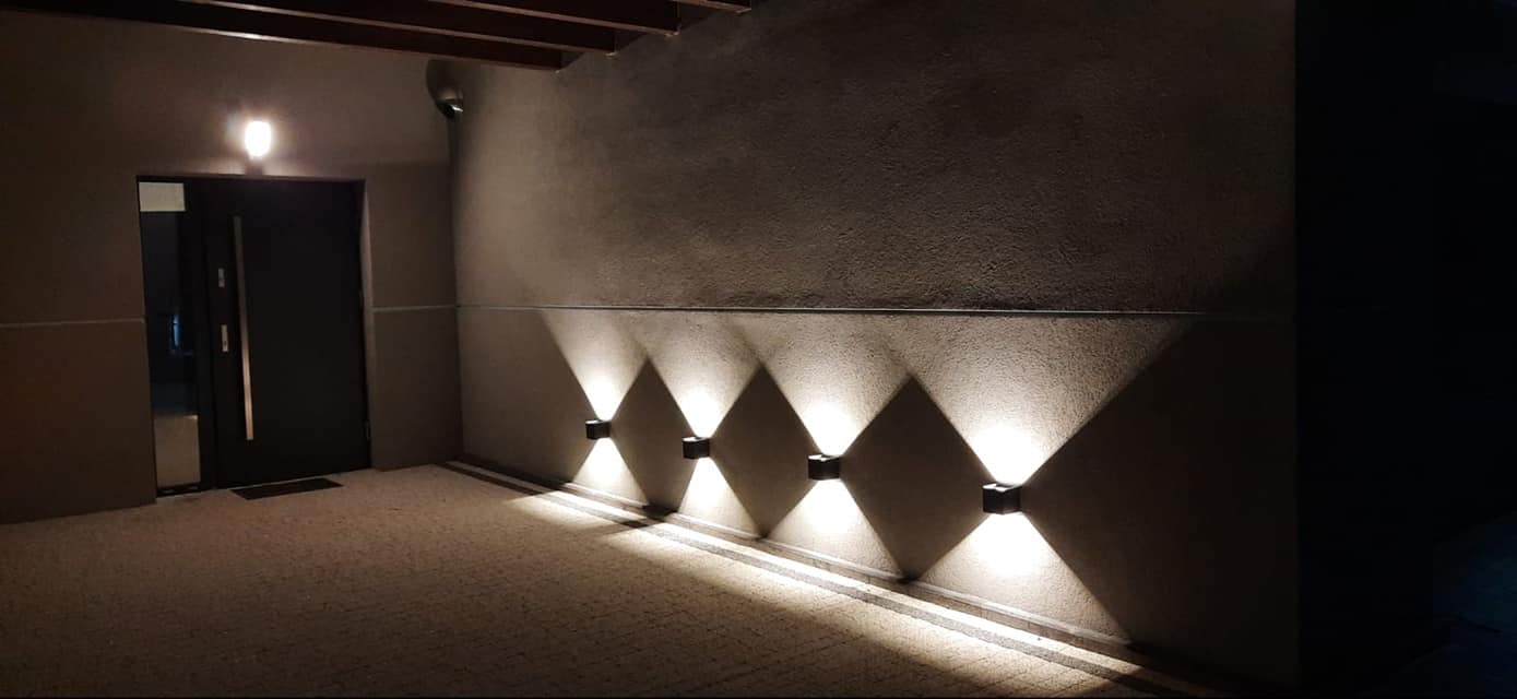 Zewnętrzne oświetlenie elewacyjne: cztery kwadratowe lampy ścienne emitują stożkowe strumienie światła na szarej ścianie obok ciemnych drzwi wejściowych.